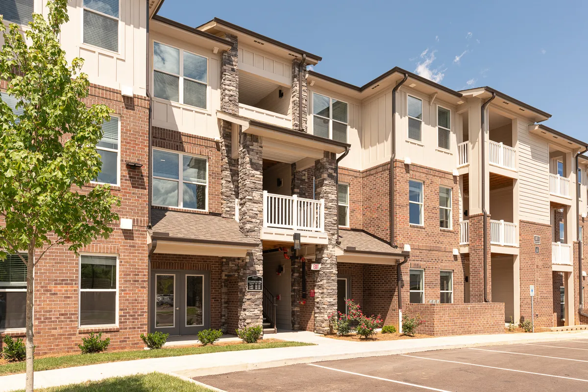 The image highlights elegant stone columns, spacious balconies, large windows, and lush landscaped greenery. The scene is bright and inviting, with residents relaxing on balconies and enjoying the outdoors.
