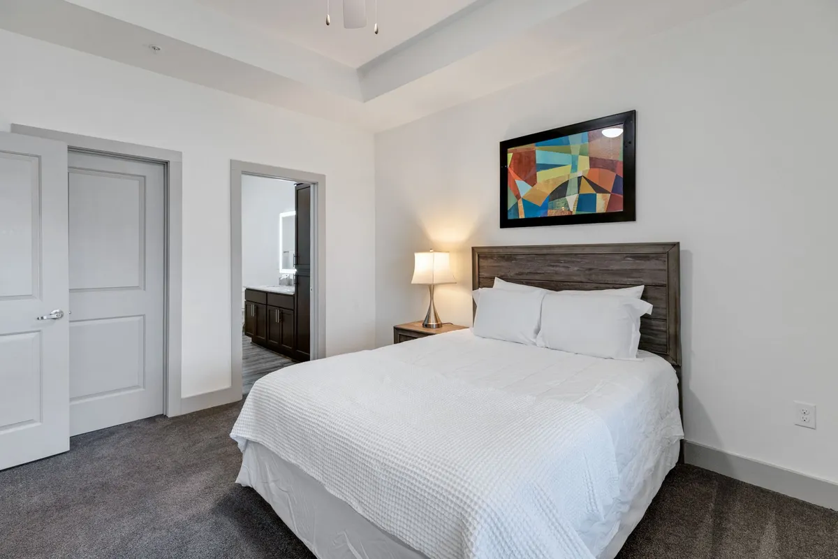 A peaceful bedroom setting with a neatly made bed, stylish lamp, creative wall art, and a doorway that hints at ample space and storage. The scene radiates a sense of calm and quiet, reflecting an ideal environment for restful sleep and rejuvenation.