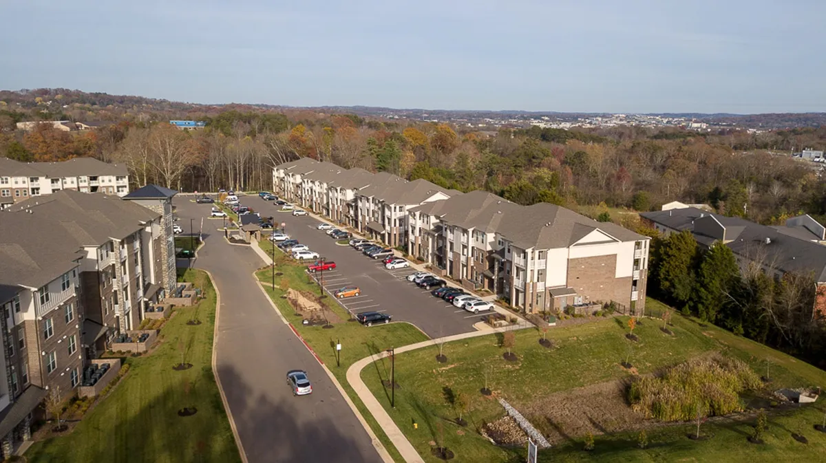 A view of the apartment buildings surrounded by landscaped green spaces, a parking lot, and mature trees. Residents are seen walking and spending time in the outdoor communal areas, evoking a sense of community and belonging.