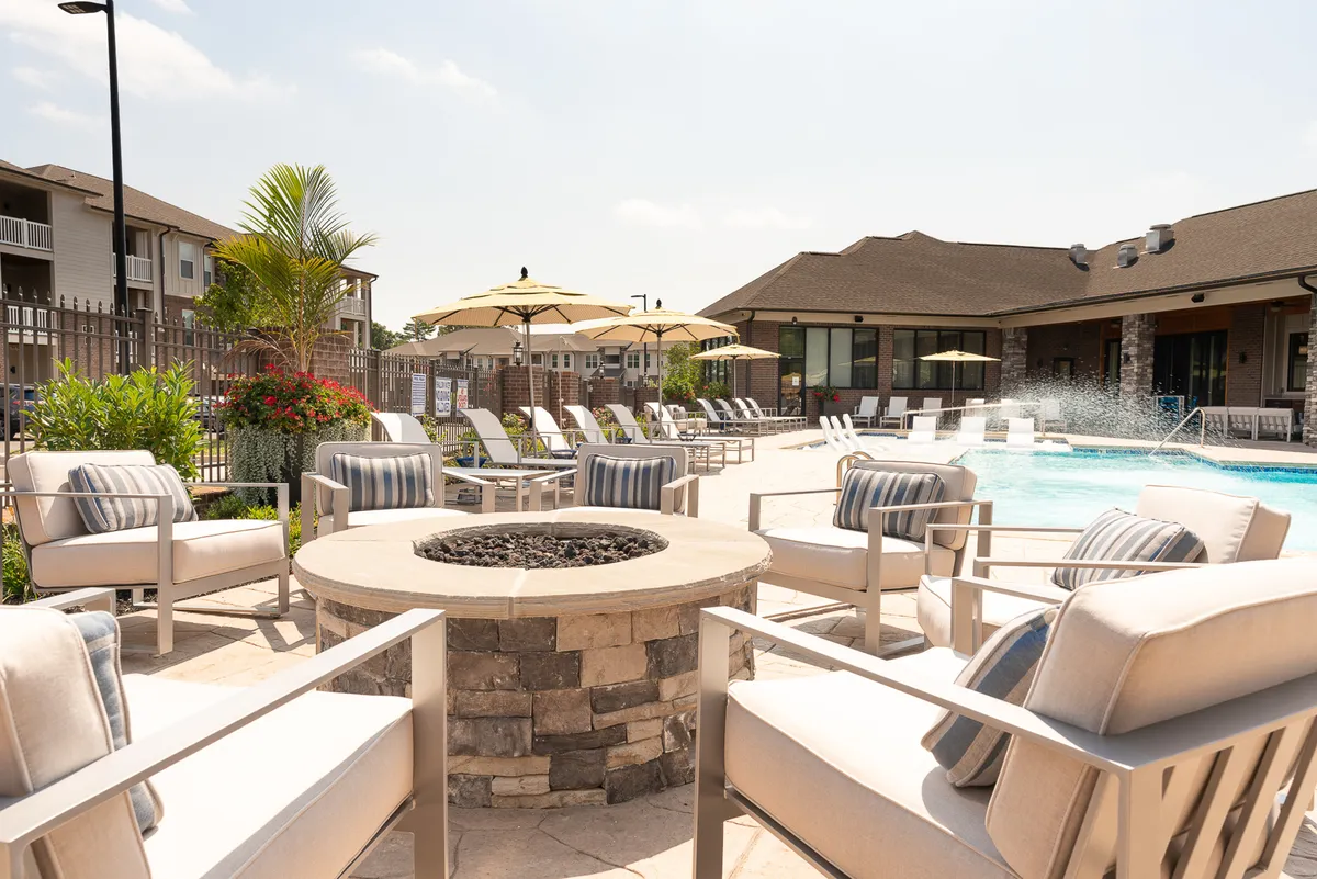 A vibrant pool area bordered by lounge chairs, umbrellas, a cozy fire pit, and manicured landscaping, all basking in natural light. The atmosphere is lively yet relaxed, ideal for relaxation or socializing day or night.