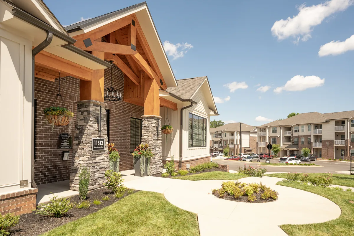 A grand entrance framed by sturdy wooden beams and a stone facade, enhanced with decorative planters and lush landscaping. The welcoming entryway sets the tone for a community-focused and accessible living environment.