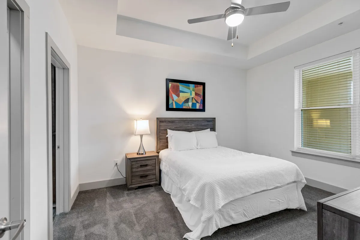 A serene bedroom featuring a comfortable bed adorned with luxurious linens, a cozy lamp on the nightstand, calming artwork above the headboard, and a large window allowing soft daylight to filter into the room. The overall atmosphere is peaceful and restful, perfect for relaxation or reading.